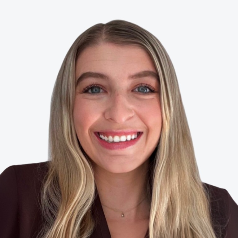 A young woman with long blond hair and blue eyes smiles at the camera. She is wearing a dark-colored blazer and standing against a plain, light background.