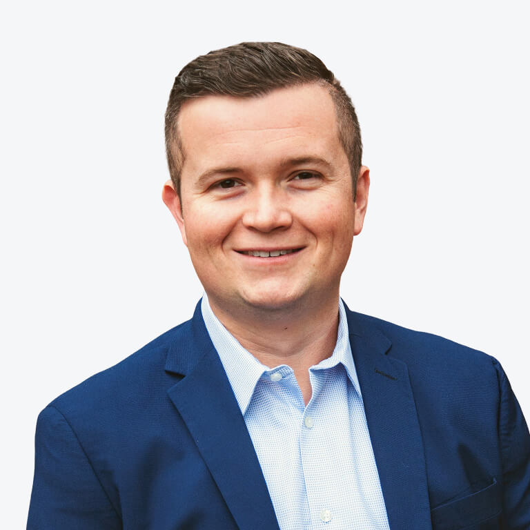 A man with short brown hair wearing a blue blazer and a light blue shirt, smiling at the camera against a plain white background.