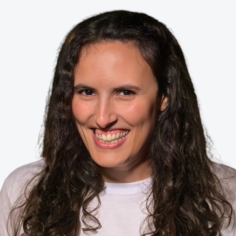 A person with long, curly, dark hair and a broad smile is wearing a white shirt, posing in front of a plain, light background.