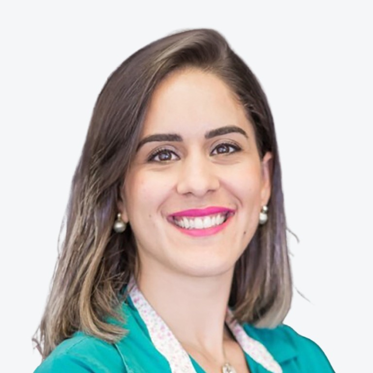 A woman with straight, shoulder-length brown hair and pearl earrings smiles at the camera. She wears a teal top with a white collar and pink lipstick. The background is plain and light-colored.