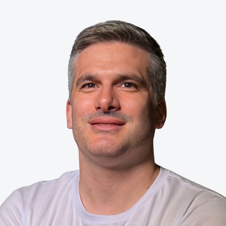 A man with short gray hair and light skin smiles slightly while wearing a white t-shirt. The background is plain and light-colored.